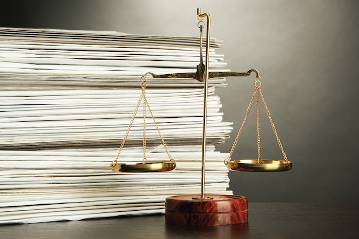 A gold scale of justice sits on a desk in front of a tall stack of papers