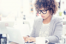 A professionally dressed woman wearing glasses with curly brown hair is typing on her computer.