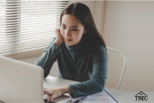 A woman seated in front of a white laptop. She's smiling while staring at the screen and wearing a green long-sleeved top. A window covered with open blinds is to her right.